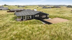 Une maison moderne dotée d’une grande terrasse, entourée d’herbes hautes et d’autres maisons, le tout situé dans un cadre rural.