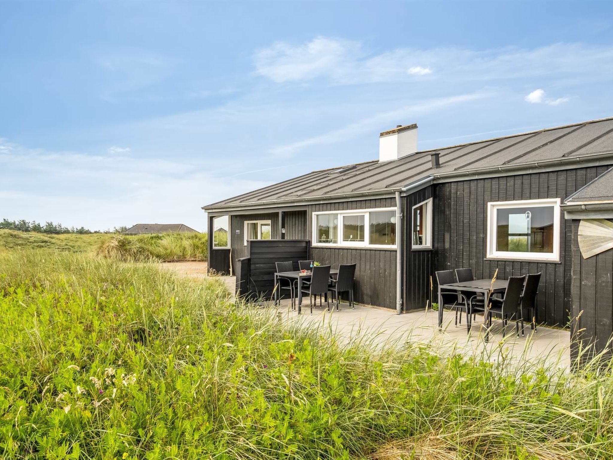 Une maison moderne, dotée d’une façade sombre, de grandes fenêtres et d’un patio aménagé avec du mobilier d’extérieur.