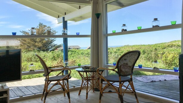 Quarto com televisão, duas cadeiras de vime, uma mesa pequena e janelas amplas com vista para vegetação e uma paisagem distante.