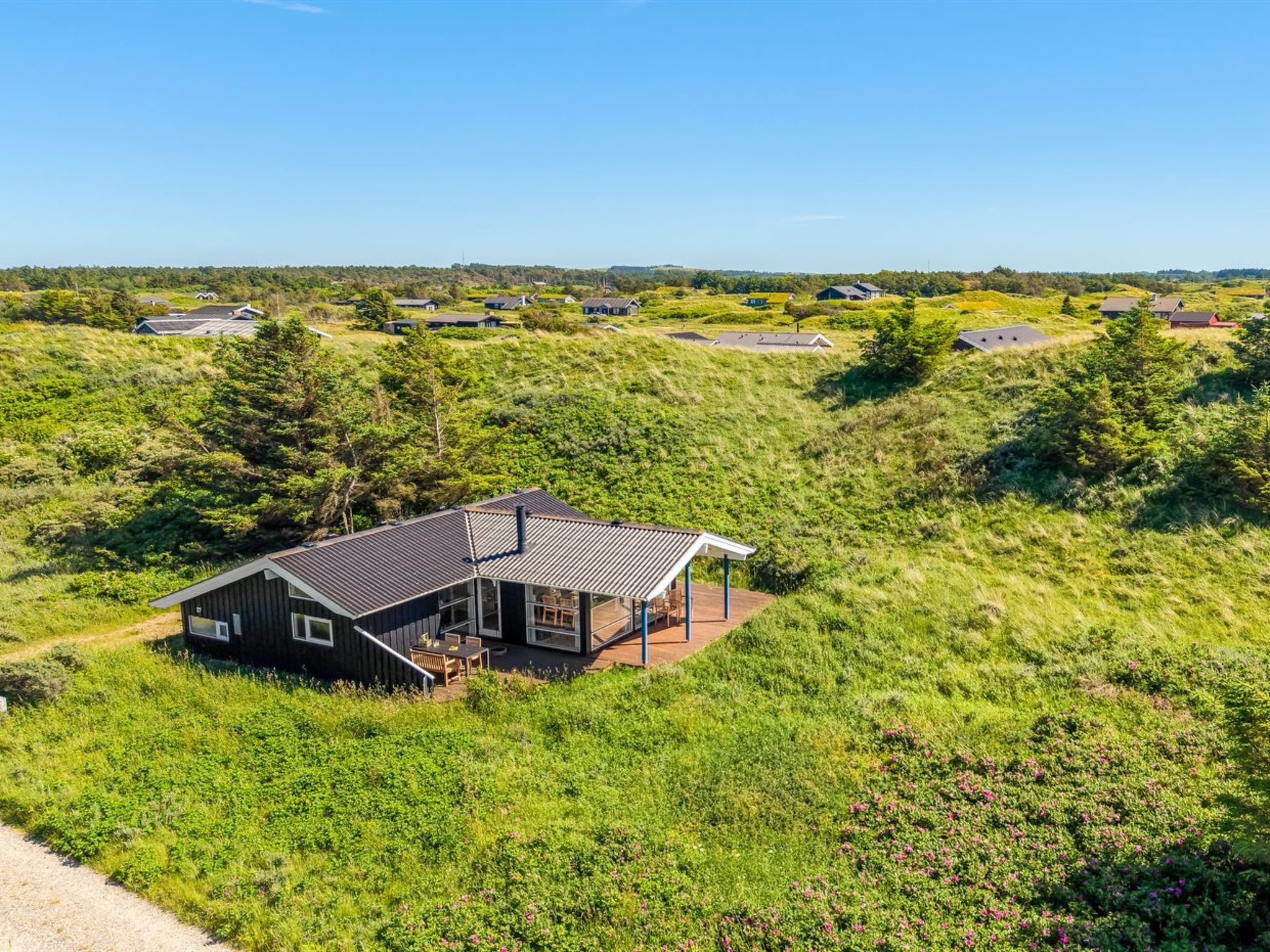Une maison moderne dotée d’une grande terrasse, entourée d’une végétation luxuriante et bordée d’autres maisons au loin.