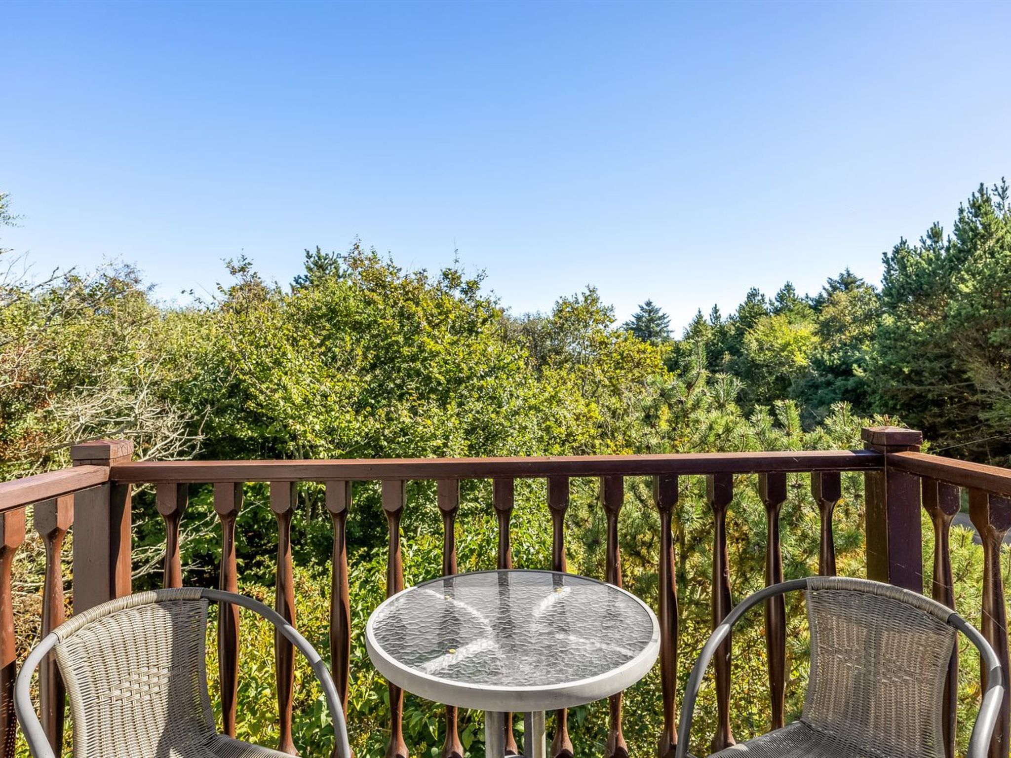 Ein Balkon mit rundem Tisch und zwei Korbstühlen, umgeben von einem Holgitter, mit Blick auf einen dichten Wald.
