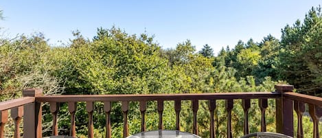 Ein Balkon mit rundem Tisch und zwei Korbstühlen, umgeben von einem Holgitter, mit Blick auf einen dichten Wald.