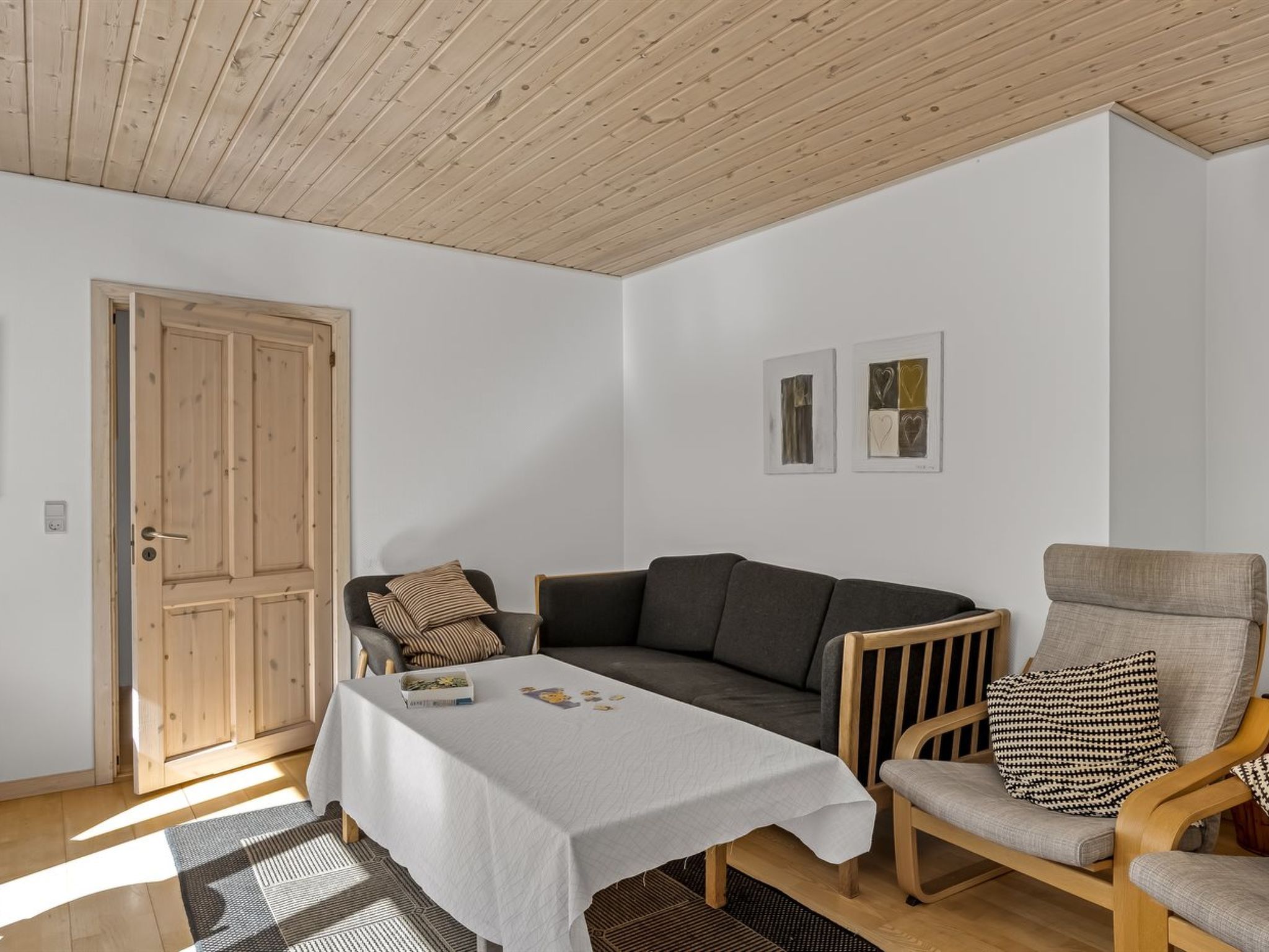 Una habitación con puerta de madera, un sofá gris, un sillón de madera, una mesita con mantel blanco y dos cuadros en la pared.