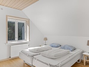 Un dormitorio con techo inclinado, ventana con vistas a árboles, cama con ropa blanca y mesita de noche con lámpara.