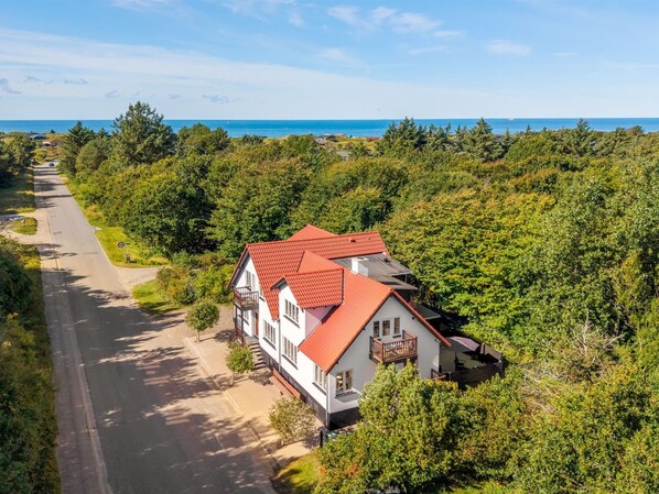 Una casa de dos pisos con techo rojo, rodeada de vegetación exuberante y una vista despejada al mar.