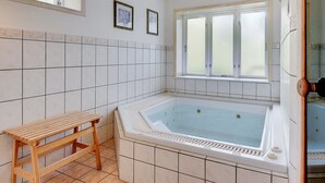 Ein Badezimmer mit Badewanne, einer Holzbank, gefliesten Wänden und zwei Fenstern.