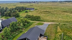 A modern house with a large deck and outdoor seating area, surrounded by a grassy field and a body of water in the background.