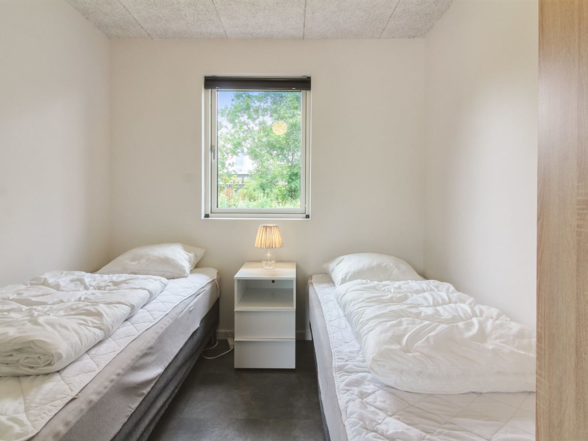 A small, single-room with two beds, a window with a view of greenery, and a bedside table with a lamp.
