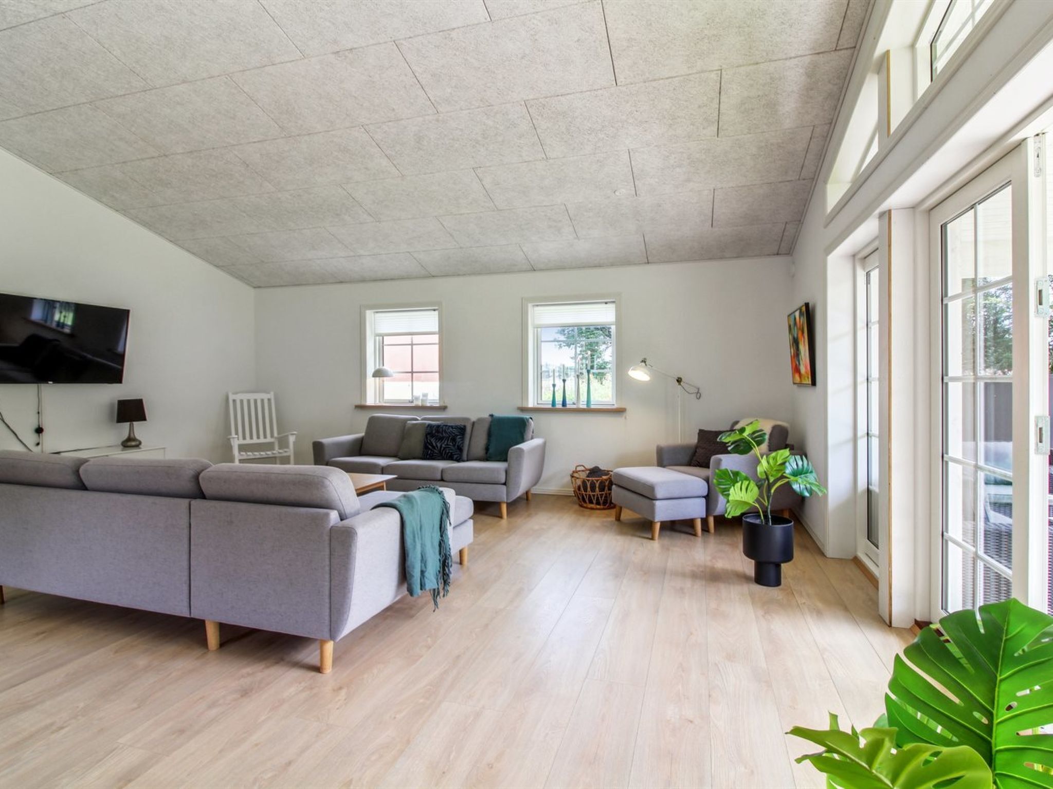 A spacious living room with a large grey sectional sofa, a smaller grey sofa, and a grey ottoman. There are two windows providing natural light, a flat-screen TV mounted on the wall, and a potted plant in the corner.