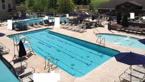 A swimming pool with multiple lanes, surrounded by lounge chairs and umbrellas, set against a backdrop of a green landscape and a building.