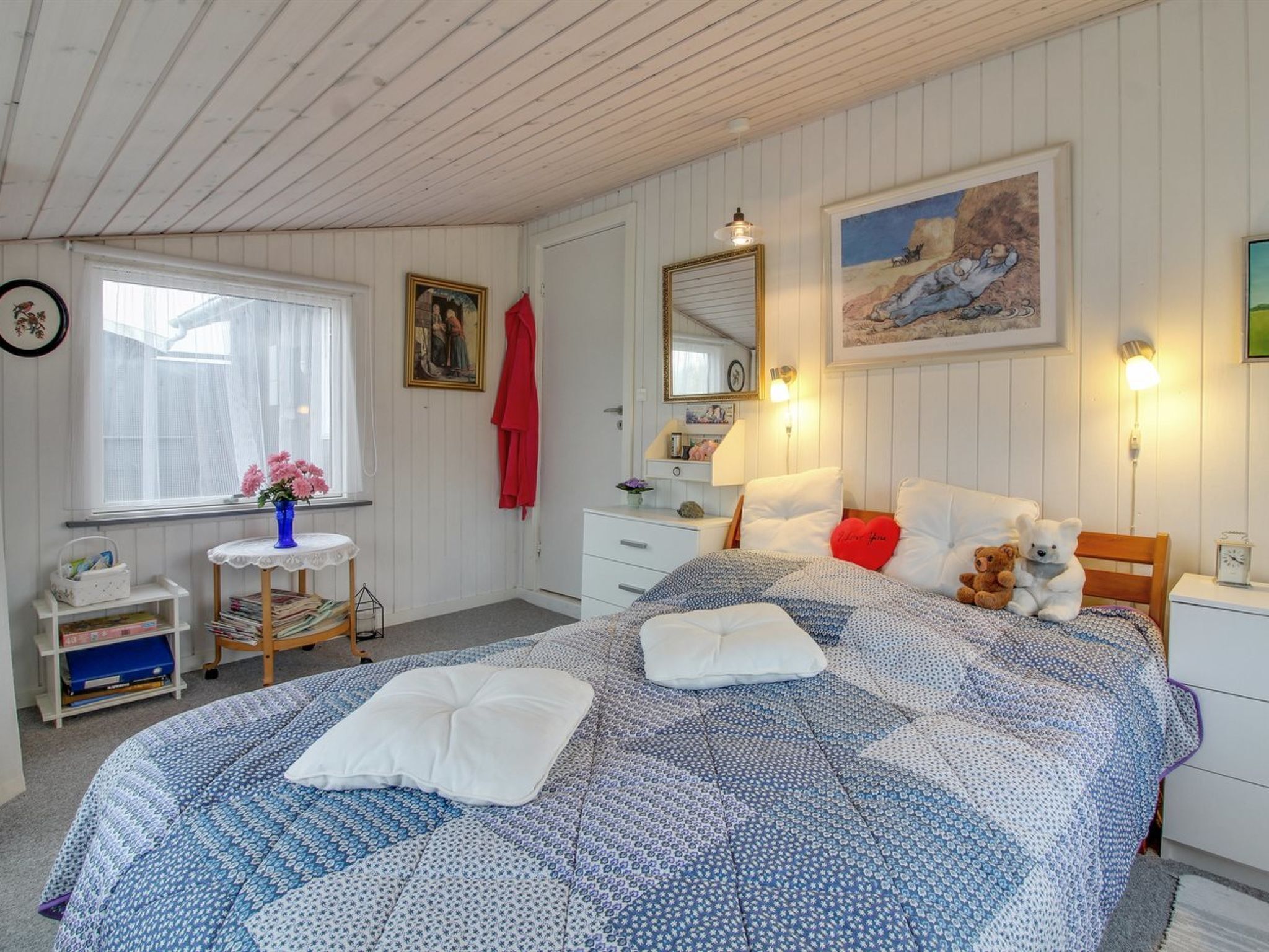 A cozy bedroom with a large bed, a bedside table, a dresser, a window with curtains, and a painting on the wall.