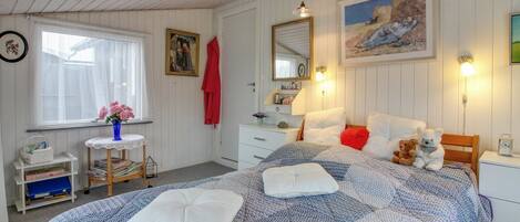 A cozy bedroom with a large bed, a bedside table, a dresser, a window with curtains, and a painting on the wall.