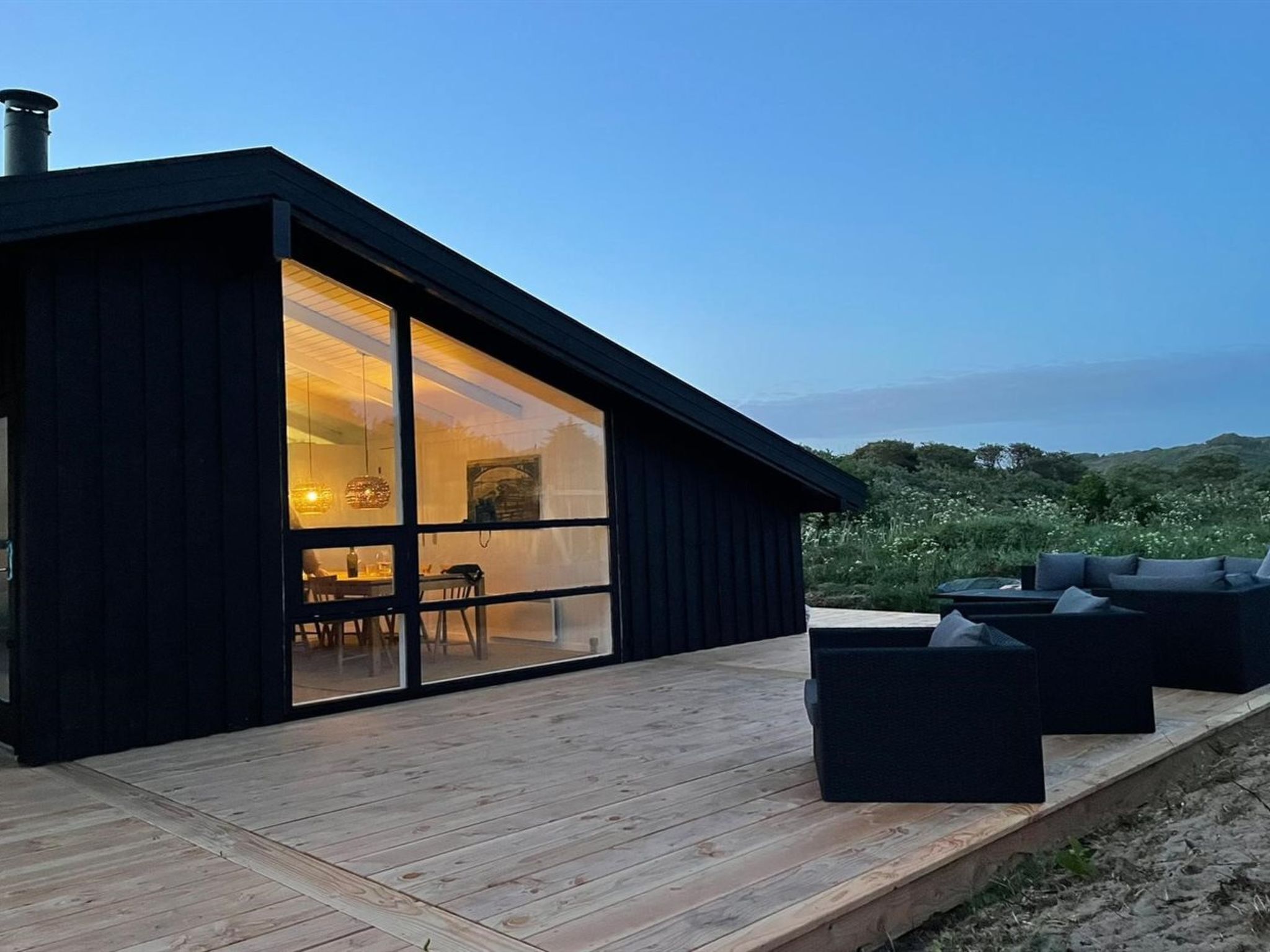 A modern house with a large glass door leading to an interior dining area, a deck with outdoor seating, and a view of the landscape.