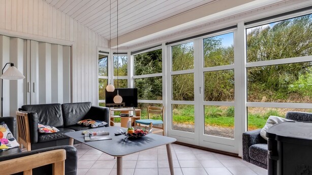 A modern living room with a black leather sofa, a wooden coffee table with fruits, and a TV. Large windows offer a view of greenery outside. - "Hauke" - 478m from the sea by Interhome (Hjørring)