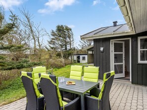 A patio with a table and chairs, a small building with a door, and a garden with trees and shrubs.
