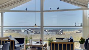 Een ruime woonkamer met grote ramen, een houten plafond en uitzicht op huizen en de zee.