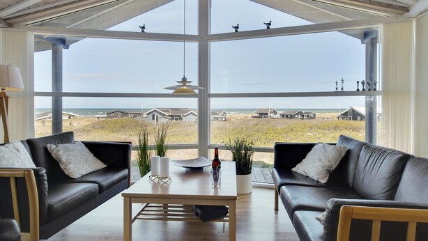 Een ruime woonkamer met een groot erkerraam met uitzicht op een strand en huizen.