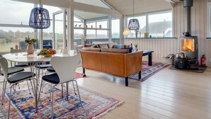 Een ruime woonkamer met een groot raam, een bank, een eettafel met stoelen en een houtkachel.