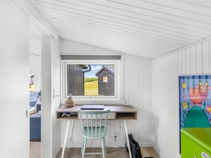 A room with a desk, a window with a view of a wooden structure, a colorful painting, and a green storage box.