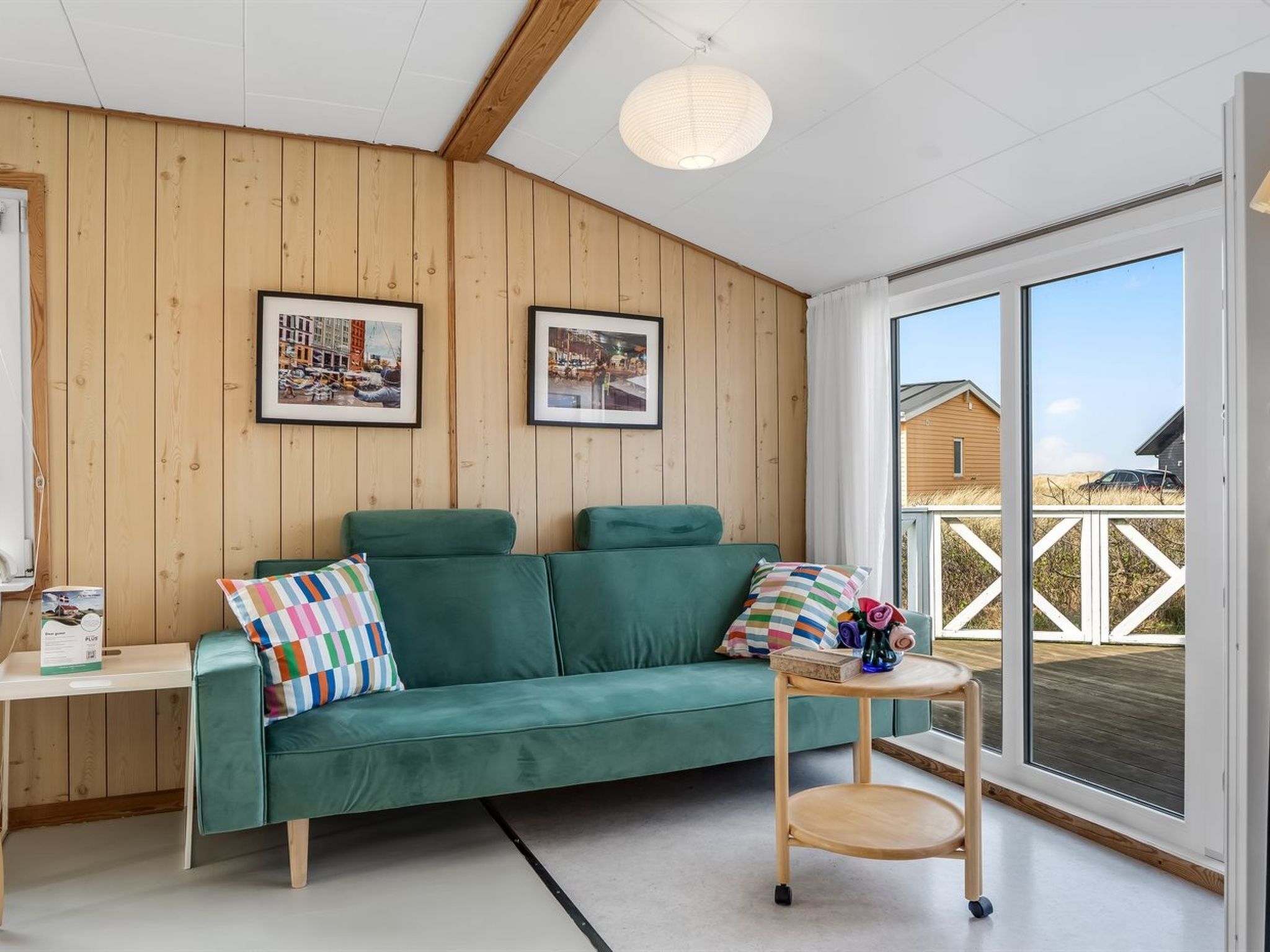 Une pièce chaleureuse avec un canapé vert, deux tableaux encadrés au mur, une petite table avec un vase, et une vue sur une terrasse en bois à l’extérieur.