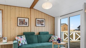 A cozy room with a green sofa, two framed pictures on the wall, a small table with a vase, and a view of a wooden deck outside.
