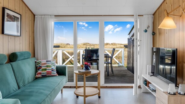 A cozy room with a teal sofa, a small wooden table, a TV, and a view of the beach through large sliding doors.