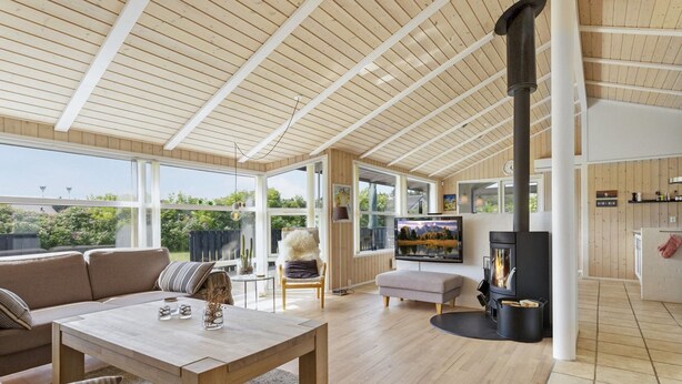 A spacious living room with a wooden ceiling, a fireplace, a television, and a large window overlooking greenery.