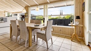 A spacious dining area with a wooden table and chairs, large windows with a view of greenery, and a small side table with a lamp.