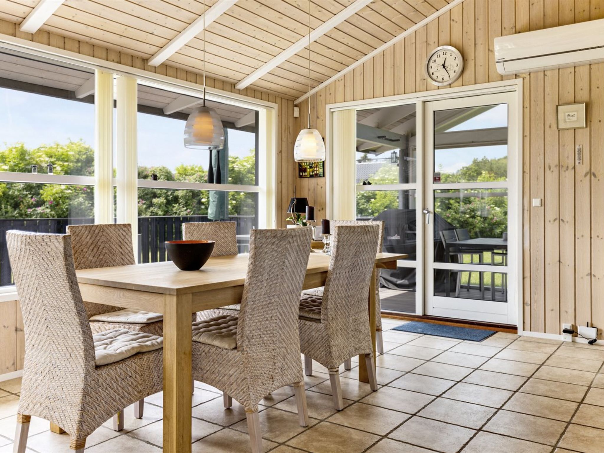 Een overdekt terras met een houten tafel en rieten stoelen, grote ramen en uitzicht op groen.