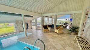 Une piscine intérieure offrant une vue dégagée sur l’extérieur, entourée de meubles en osier et dotée d’un sol carrelé.