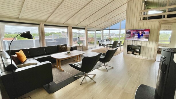 Un vaste espace de vie comprenant un canapé, une table basse et des fauteuils, de grandes fenêtres offrant une vue sur l’extérieur et un téléviseur fixé au mur.