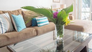 Ett vardagsrum med en beige soffa, mönstrade kuddar, ett soffbord i glas och en vas med gröna växter.