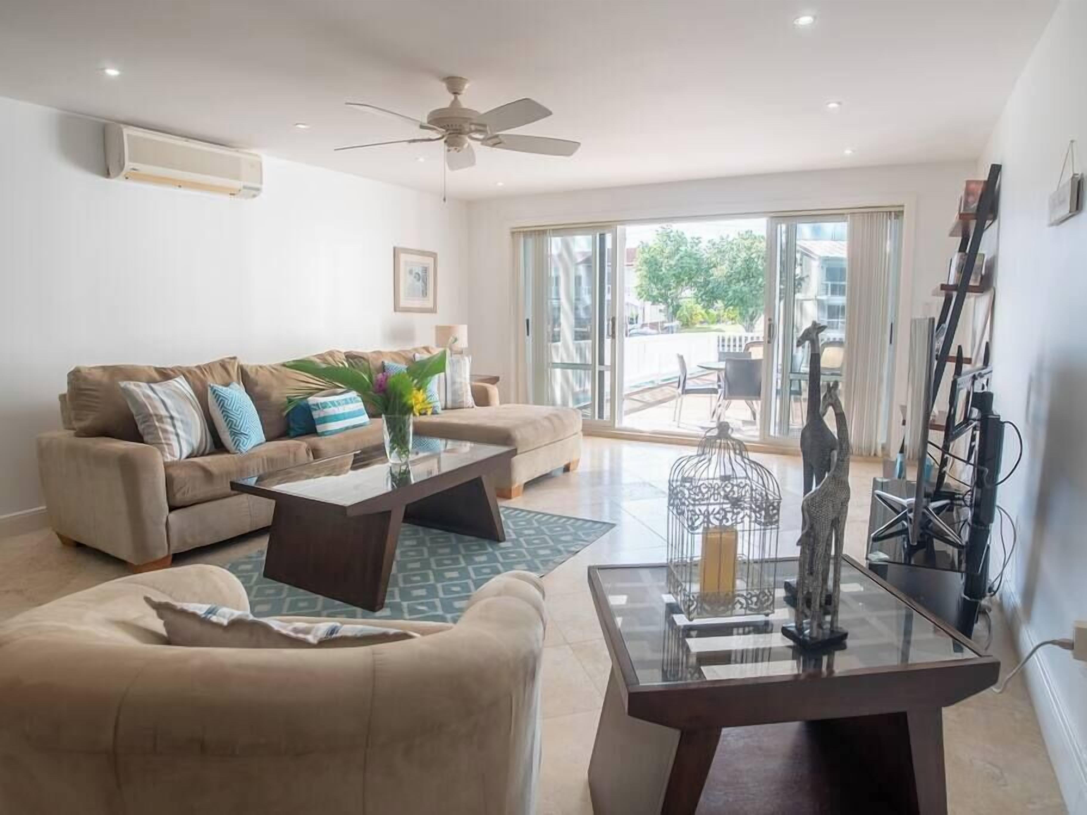 A living room with a sofa, coffee table, and decorative items. There is a ceiling fan, large windows, and a view of the outdoors.