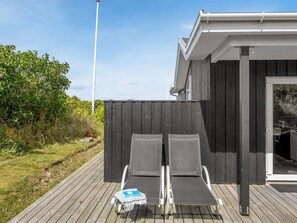 A deck with two lounge chairs and a small table, a wooden fence, and a building with a dark exterior and a white roof.