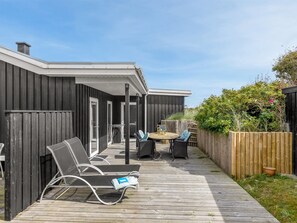 A covered outdoor deck with lounge chairs and a table, surrounded by a wooden fence and vegetation.