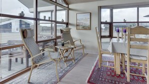 Une pièce spacieuse avec de grandes fenêtres, un plafond en bois et une vue sur l’extérieur. On y trouve deux chaises en bois, une table et un tapis.