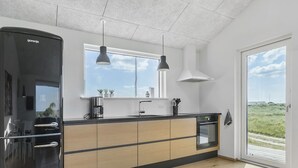 Eine moderne Küche mit einem schwarzen Kühlschrank, Holzschränken, einer Kochinsel mit Glasplatte, Pendelleuchten und Blick ins Freie.