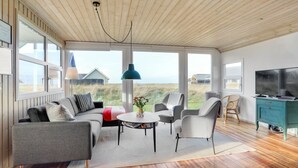 Una sala de estar moderna con techo de madera, ventanas amplias, un sofá, sillones, una mesa de centro y una televisión.