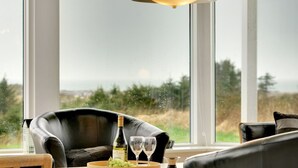 En moderne stue med et stort vindue, en lædersofa, et træbord med en vinflaske og glas, samt en pendellampe.