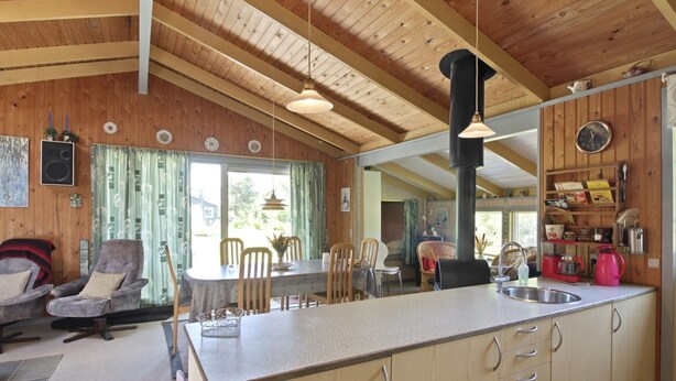 Interior acogedor de una cabaña con techo de madera, comedor con mesa y sillas, cocina con lavaplatos y armarios, y sala de estar con sofá y sillones.