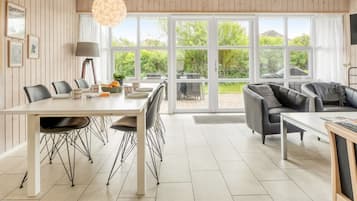 Un vaste espace de vie comprenant une table à manger, des chaises, un canapé et de grandes fenêtres donnant sur l’extérieur.