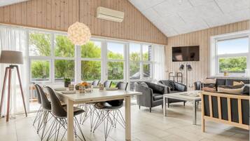 Un vaste espace de vie comprenant une table à manger, des chaises, un canapé et un téléviseur. De grandes fenêtres laissent entrer la lumière naturelle et offrent une vue sur la verdure environnante.