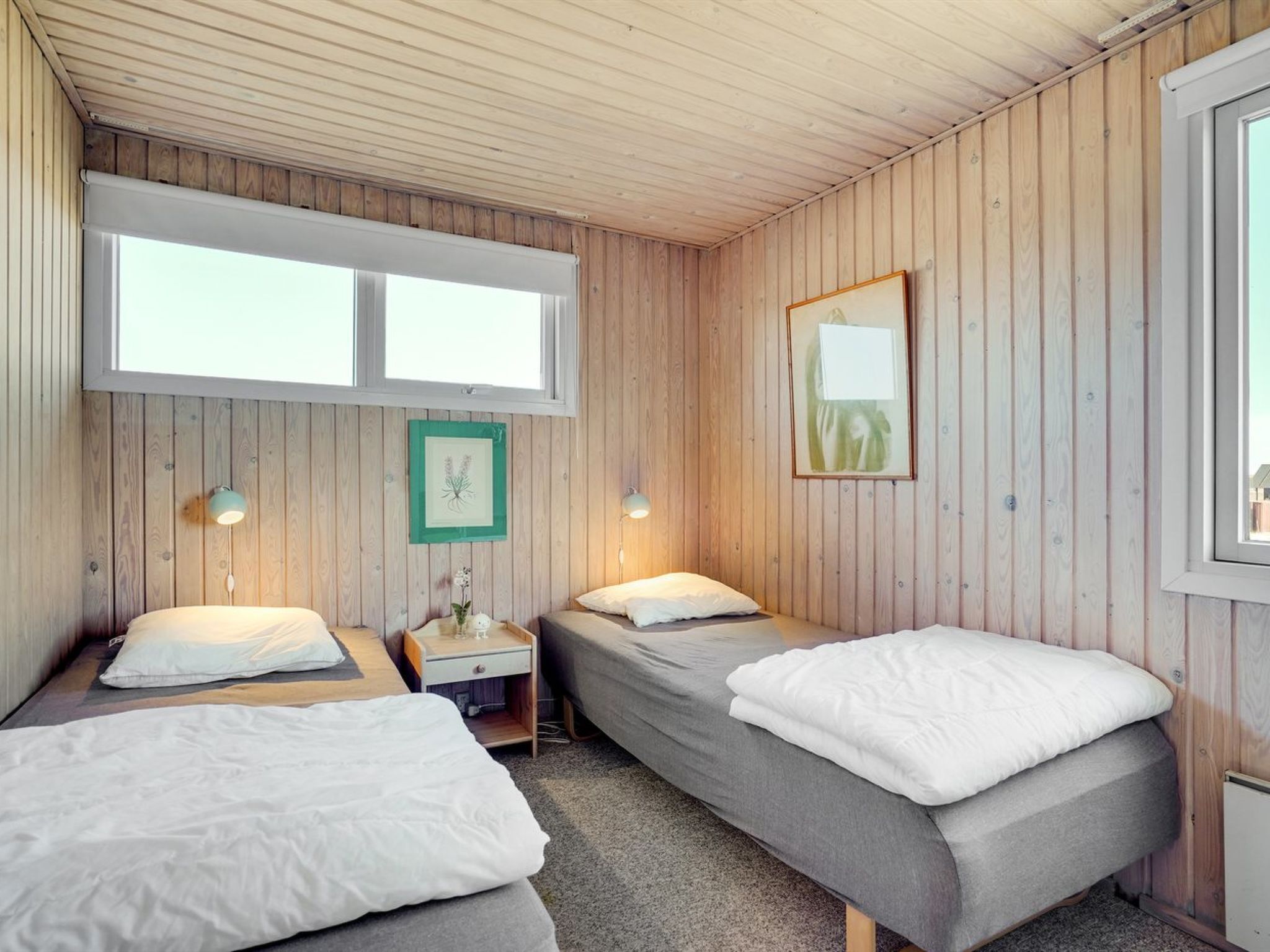 A room with two single beds, a small desk, a window, and wooden walls.