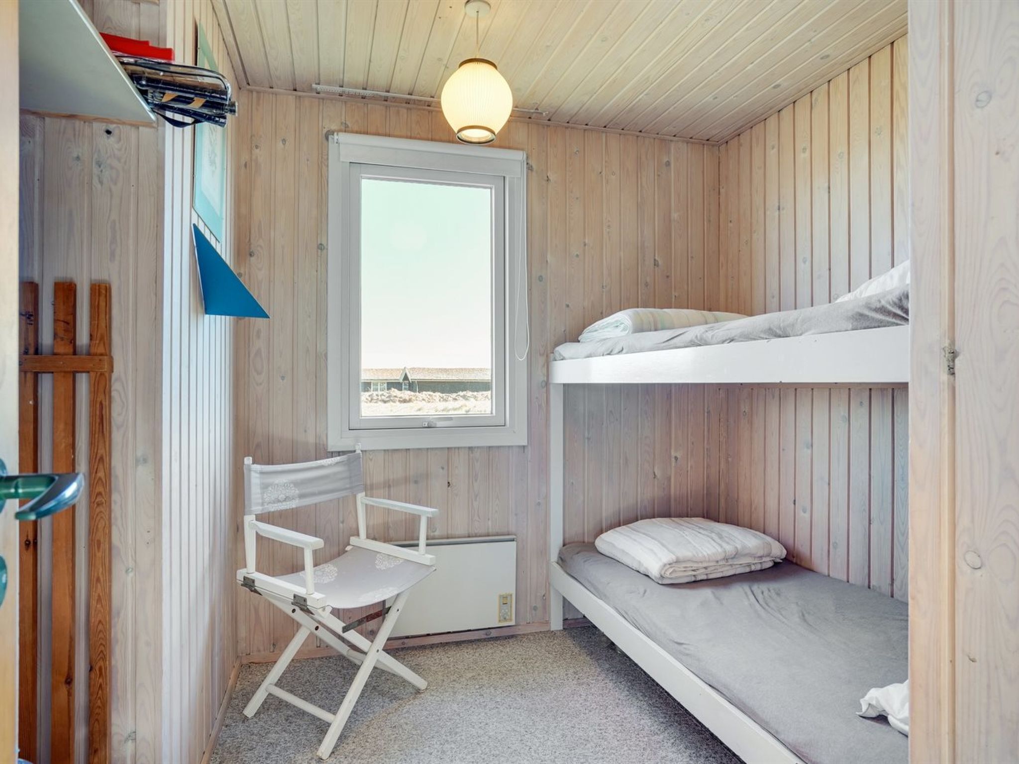 Una habitación pequeña con una cama litera, una silla plegable, un ventanal con vista al mar y una lámpara en el techo.