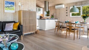 Un interior moderno con techo de madera, zona de comedor con mesa y sillas, cocina con armarios blancos y sofá negro con cojines.