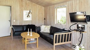 Una habitación acogedora con un sofá de cuero negro, una mesa de centro de madera, un televisor montado en la pared y una ventana con vistas a zonas verdes.