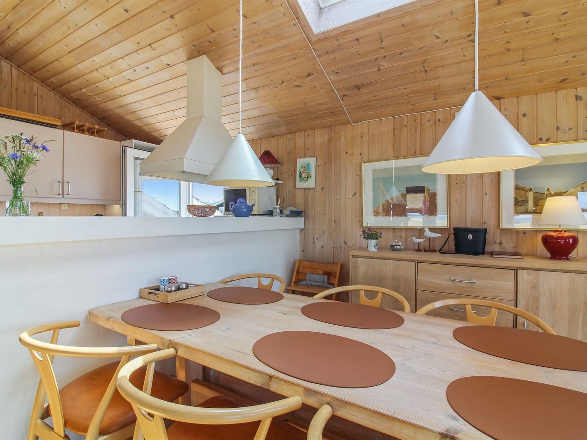 Une cuisine moderne avec des meubles en bois, une table à manger entourée de chaises marron, des luminaires suspendus et une lucarne.