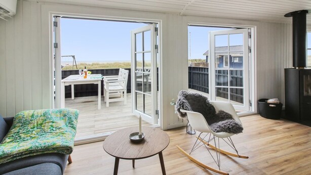 A spacious living area with a wooden floor, a sofa, a rocking chair, and a small table. There are large glass doors leading to an outdoor deck with a table and chairs. - "Adina" - 50m from the sea by Interhome (Hjørring)