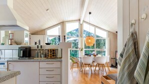 Een moderne keuken met witte kasten, een inbouwoven en een koffieapparaat. Er is een eethoek met een tafel en stoelen, en een groot raam met uitzicht op de omgeving.