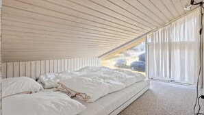 Un dormitorio con techo inclinado, cama con ropa de cama blanca y vistas al exterior a través de una ventana con cortinas traslúcidas.